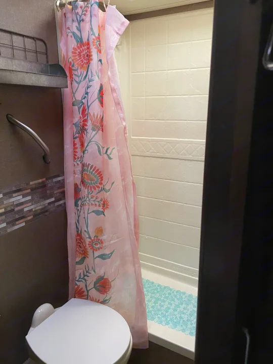 This RV rental bathroom features a floral shower curtain, toilet, and compact shower area with a blue mat.. Thor Four Winds 2018