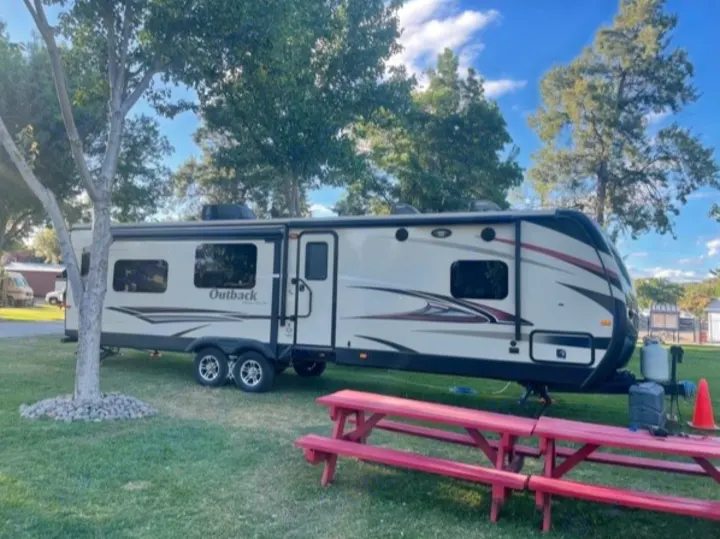 Modern tan Outback travel trailer RV rental at a green campsite with a red picnic table under a sunny sky.. Keystone 316-UL 2014