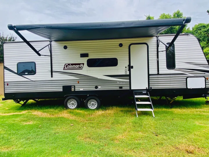 Side view of a white and grey Coleman travel trailer with its black awning extended, set on green grass. Great for RV rental.. Coleman Lantern 2021