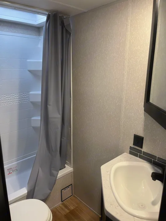 A full view of a clean, modern RV rental bathroom featuring a shower with a gray curtain, toilet, and sink.. Forest River Cherokee Grey Wolf Special Edition 2022