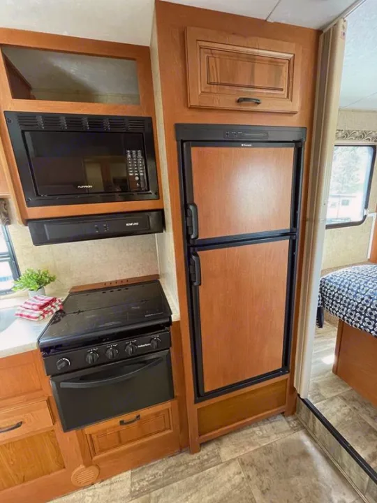 View of a functional RV rental kitchen with light wood cabinetry and integrated black microwave, stove, oven, and refrigerator.. Sunseeker 2650 2015