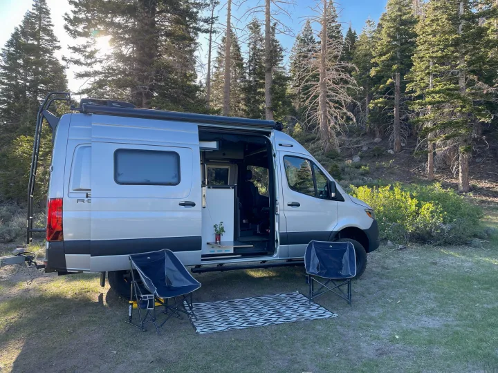 RV rental: Silver camper van at a peaceful wooded campsite, with chairs and mat set up outside for relaxation.. Winnebago Revel 2022
