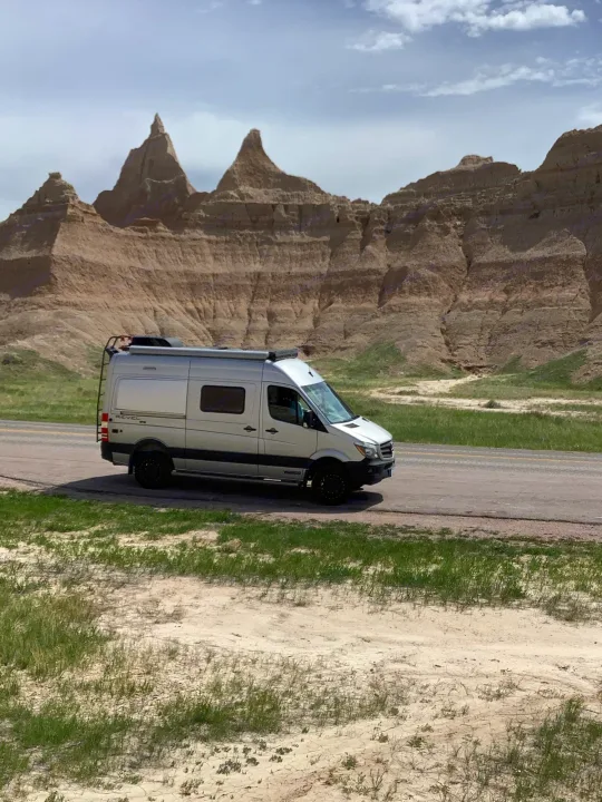 A silver RV rental camper van drives past the unique rock formations of Badlands National Park under a cloudy sky.. Winnebago Revel 2019
