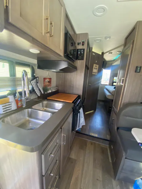 Modern RV rental interior showing a kitchen with double sinks, microwave, and ample storage, extending to a living area.. Winnebago Minnie Winnie 2020