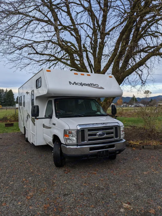 This white Majestic RV rental is parked on gravel under a large, mossy tree, with distant hills in the background.. Thor Four Winds 2013