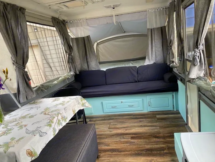 Cozy rv rental pop-up camper interior with dark sofa, light blue cabinetry, wood flooring, and bed area in back. Grey curtains.. Coleman Grand Tour Sun Valley 2002