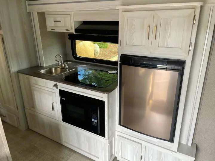 This RV rental kitchen features light wood cabinets, a sink, glass-top stovetop, built-in microwave, and a stainless steel Dometic refrigerator.. Forest River Flagstaff E-Pro 2020