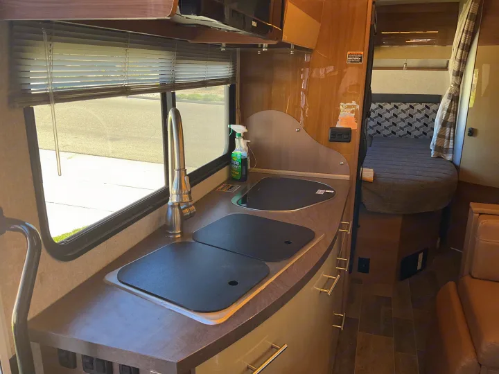Inside an RV rental: view of the kitchen galley with sink, covered stovetop, window blinds, and a glimpse of the comfy bed area.. Winnebago Navion 24J 2018