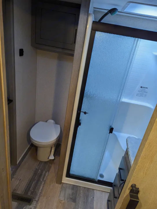 A compact rv rental bathroom featuring a white toilet, glass-enclosed shower, wood-look floor, and storage cabinet.. Keystone Raptor 2021