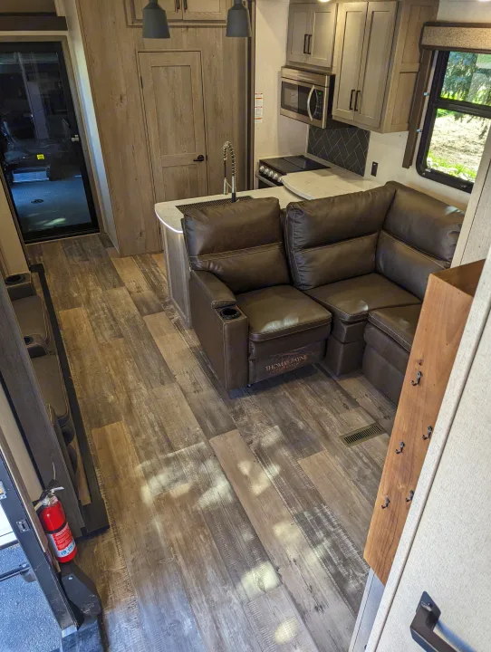 Modern RV rental interior: brown sectional sofa, full kitchen, and wood-look flooring, with a rear utility door visible.. Keystone Raptor 2021