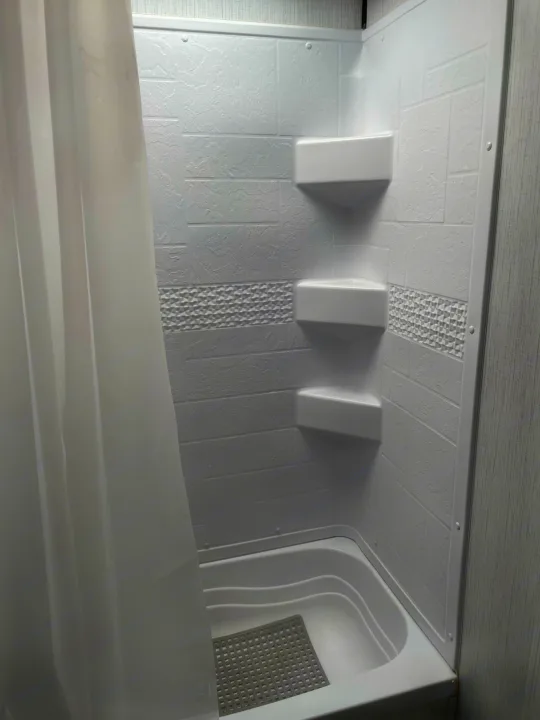 RV rental shower/tub with white textured walls, three corner shelves, a shower curtain, and bath mat.. Heartland Prowler 2021