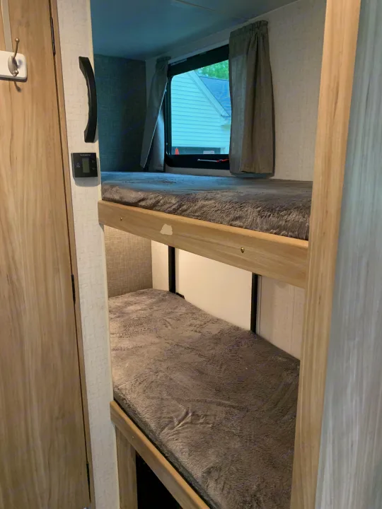 An RV rental interior showing two stacked bunk beds with grey blankets. A window is above the top bunk, with a control panel nearby.. Winnebago Micro Minnie 2021