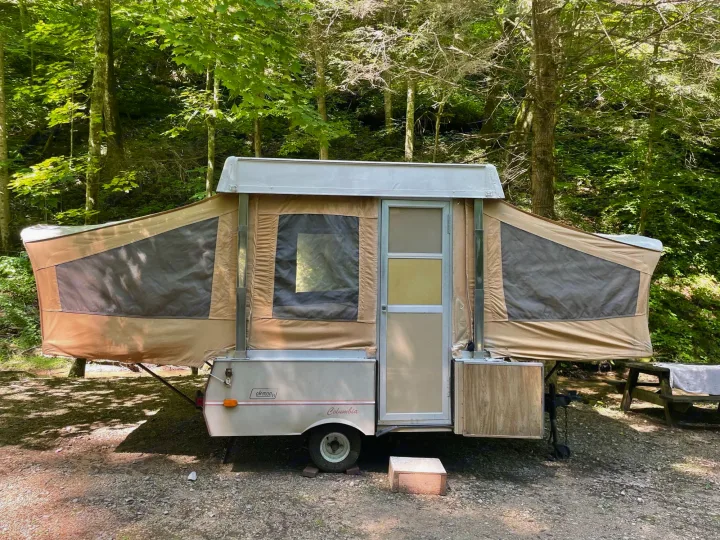 Tan pop-up camper, an RV rental, parked on gravel in a lush forest campsite. Ideal for outdoor adventures.. Coleman Other 1983