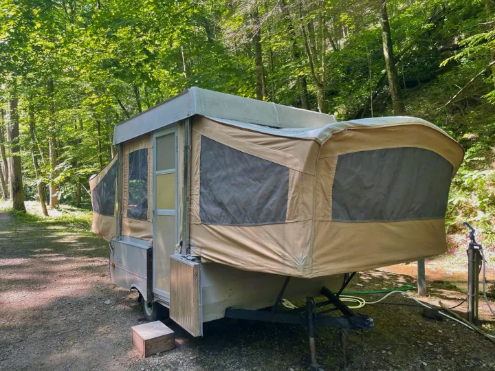 A vintage pop-up camper is set up in a wooded campsite, ideal for an RV rental adventure.. Coleman Other 1983
