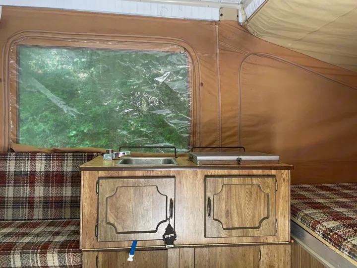 Inside a cozy pop-up RV rental: vintage kitchen with sink, covered stove, and classic plaid sleeping areas. Green view outside.. Coleman Other 1983