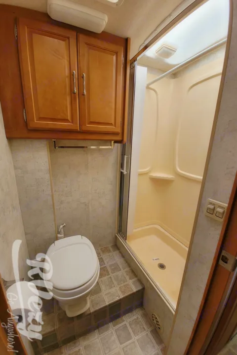 Inside an RV rental: a clean white toilet, compact shower area with sliding door, and wooden storage cabinets.. Winnebago Chalet 2007