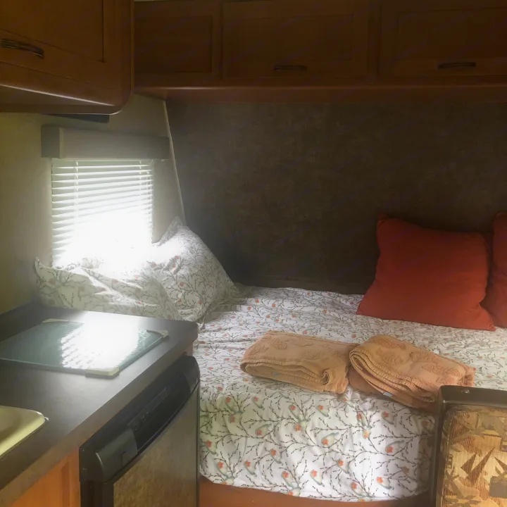 Inside an RV rental: a cozy bedroom with bird-patterned bedding, rust pillows, and a small kitchen area with cabinets.. Forest River Cherokee 2014