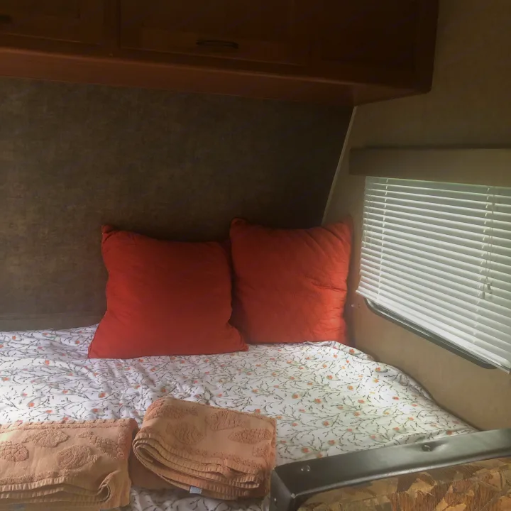 RV rental bedroom with a bed, floral sheets, orange pillows, folded towels, overhead cabinets, and a window with blinds.. Forest River Cherokee 2014
