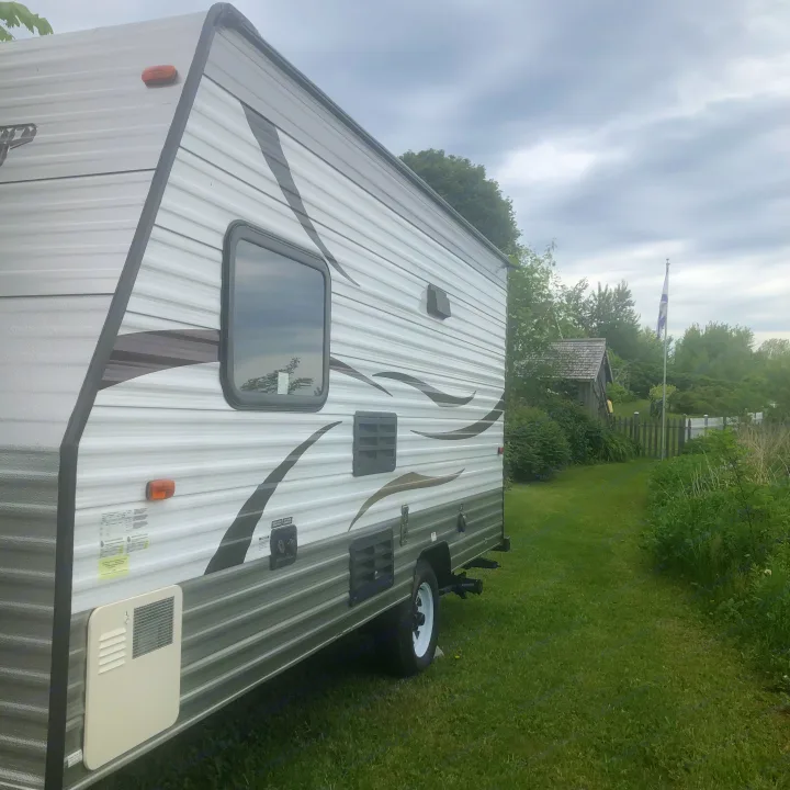 A white and grey RV rental, a travel trailer, is parked on green grass, with a shed and Quebec flag in the cloudy distance.. Forest River Cherokee 2014