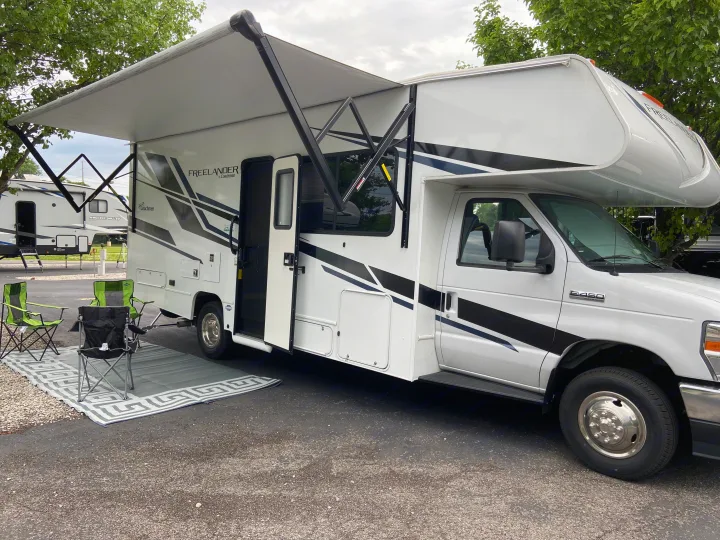 White RV rental (Coachmen Freelander) parked with awning extended. Outdoor chairs and mat create a cozy campsite setup.. Forest River Coachmen Freelander Premier 2022