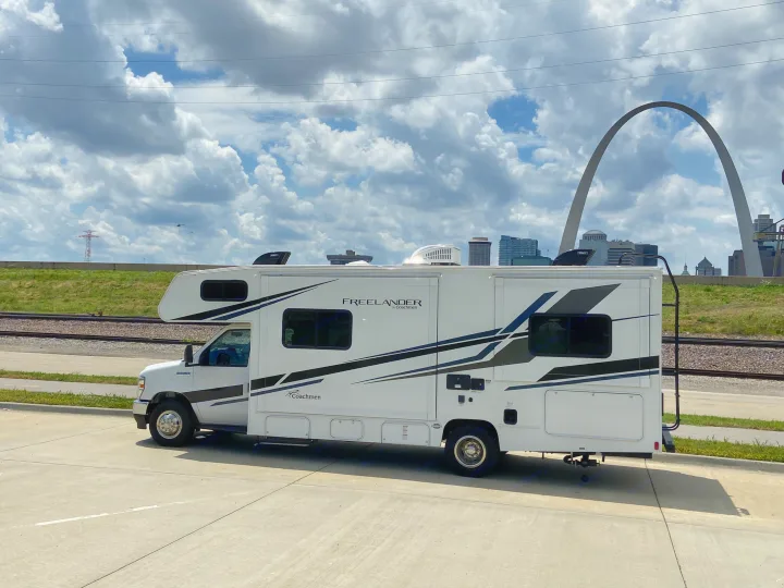 An RV rental, white with black stripes, parked on pavement, with the St. Louis Gateway Arch and city skyline under clouds.. Forest River Coachmen Freelander Premier 2022