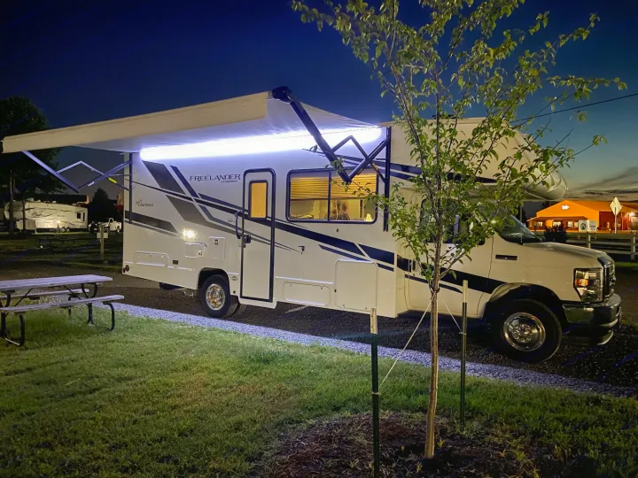 Nighttime view of an RV rental with its awning extended and lights on, parked at a campsite with a picnic table.. Forest River Coachmen Freelander Premier 2022
