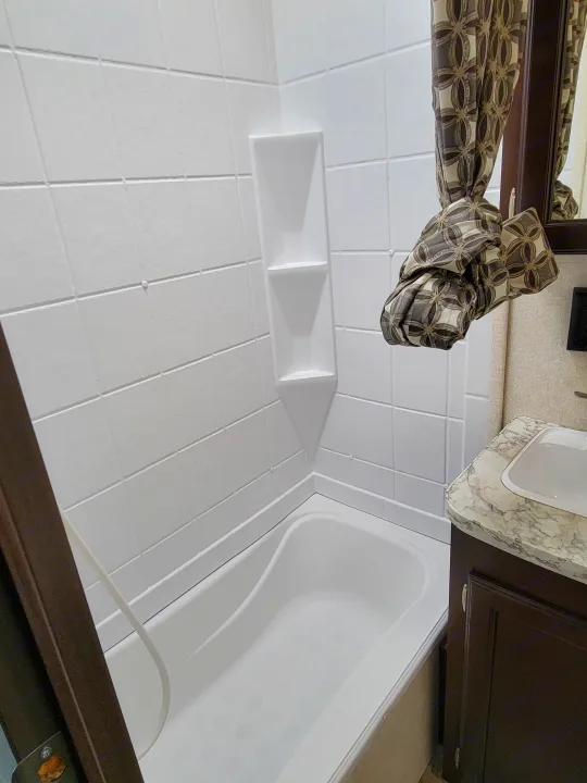 Clean white tiled shower and tub area with built-in shelves in an RV rental bathroom, featuring a decorative patterned curtain.. Jayco Jay Flight 2020