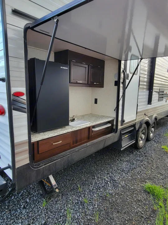 RV rental's outdoor kitchen with open panel, showing fridge, sink, and cabinets. A travel trailer on gravel.. Jayco Jay Flight 2020