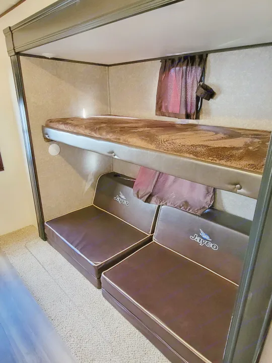 Inside an RV rental, a brown bunk bed and dark Jayco sofa with white piping create a cozy sleeping/living space.. Jayco Jay Flight 2020