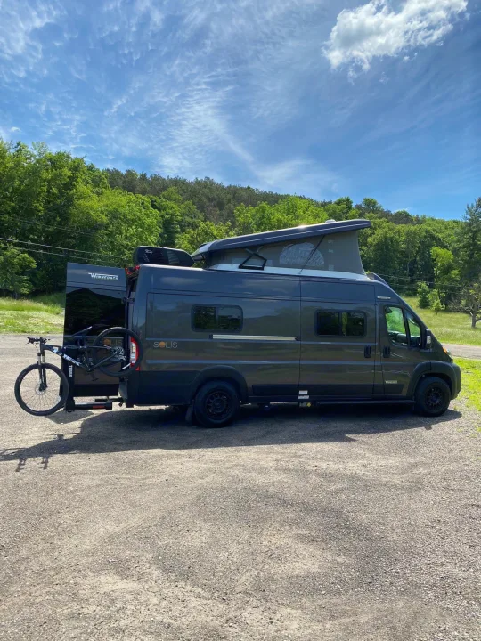 Winnebago Solis RV rental ready for adventure: dark gray van with pop-top, bike rack, parked amidst trees.. Winnebago Solis 2022