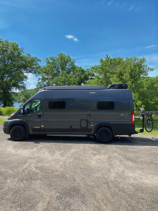 This gray Winnebago Solis RV rental van, with a bike rack, is parked on gravel near a lake and trees.. Winnebago Solis 2022