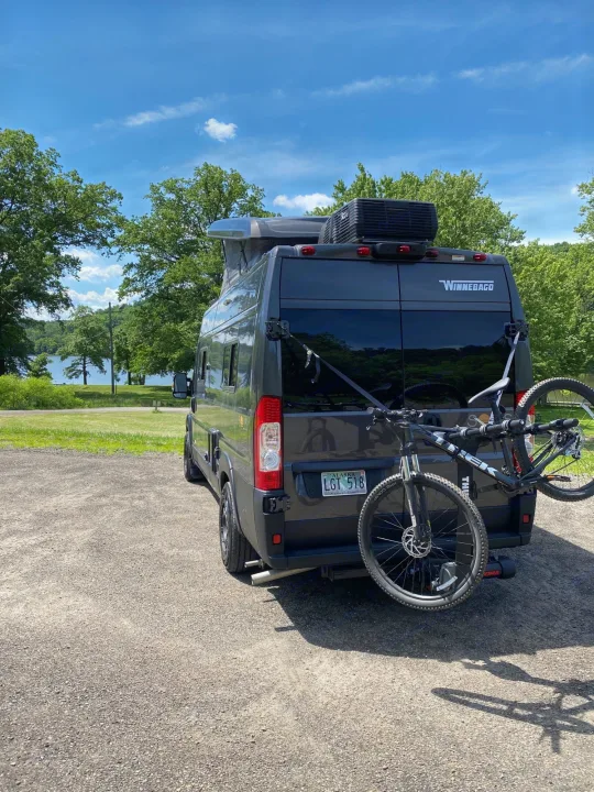 Winnebago rv rental: Dark grey camper van with a bike rack parked near a scenic lake on a sunny day.. Winnebago Solis 2022