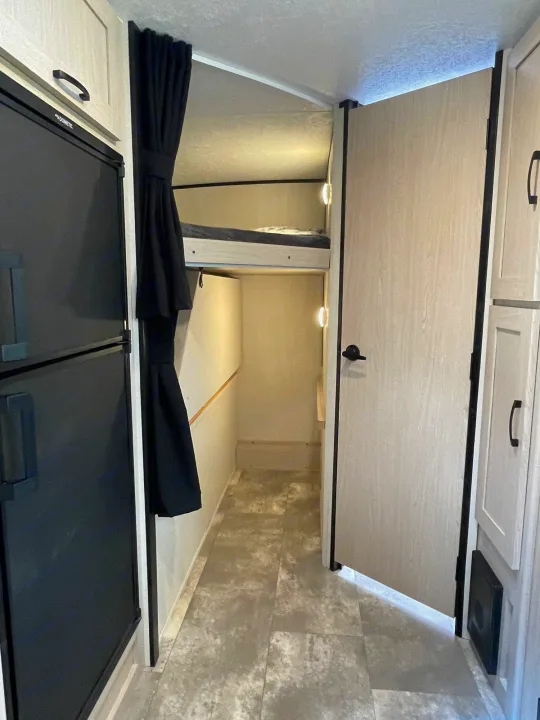 Compact RV rental interior featuring bunk beds, a black refrigerator, and a light-colored door leading further inside.. Coachmen Apex 2022