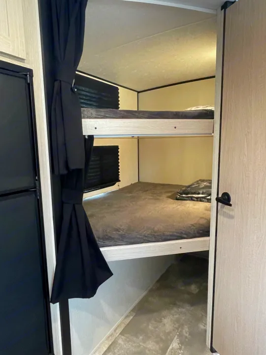 RV rental interior showcasing comfy bunk beds with privacy curtains and a compact fridge nearby.. Coachmen Apex 2022