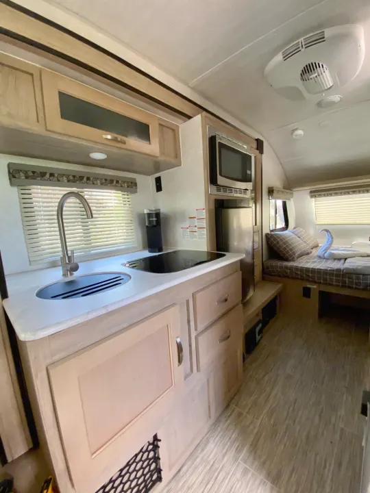 RV rental interior: compact kitchen area with sink, stovetop, microwave, fridge, and overhead cabinets. Beyond, a bed with a towel swan.. R-Pod R-Pod Trailer 2021
