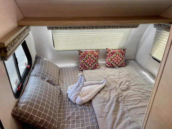 Cozy RV rental bedroom features a bed with grey plaid and plain bedding, decorative pillows, and a swan-folded towel. An emergency exit window is visible.. R-Pod R-Pod Trailer 2021