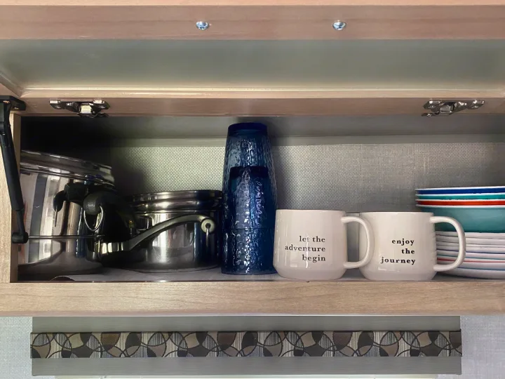 RV rental kitchen cabinet with pots, blue glasses, mugs ("let the adventure begin," "enjoy the journey"), and colorful dishes, ready for your trip.. R-Pod R-Pod Trailer 2021