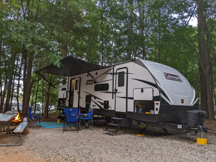 A white Coleman RV rental is parked at a gravel campsite, awning extended. A campfire burns beside a picnic table and blue chairs. Ideal for nature camping.. Dutchmen Coleman Light 2021