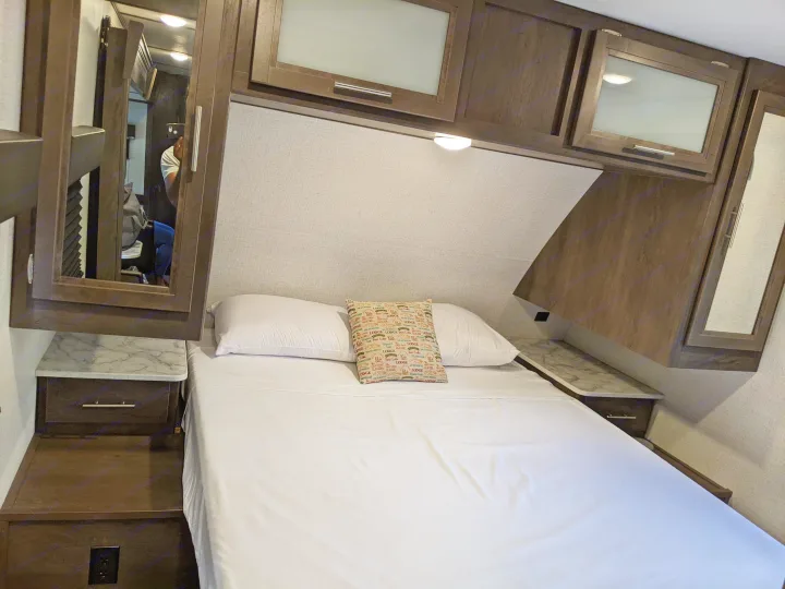 Cozy RV rental bedroom features a comfy bed with white sheets, overhead cabinets, and practical side tables.. Dutchmen Coleman Light 2021