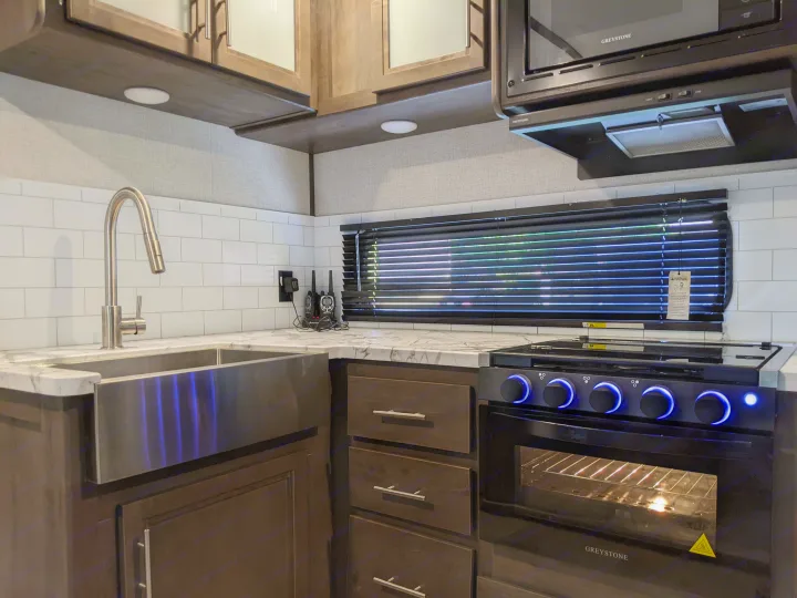 Modern RV kitchen with a stainless steel sink, glowing blue stove knobs, and subway tile, ready for your RV rental adventure.. Dutchmen Coleman Light 2021