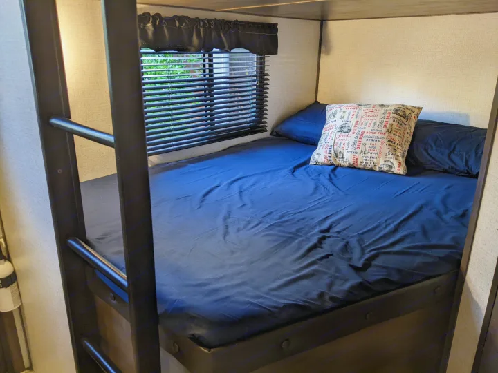 Cozy RV bunk bed with dark blue bedding, a travel pillow, and window blinds. Perfect for your next RV rental adventure.. Dutchmen Coleman Light 2021