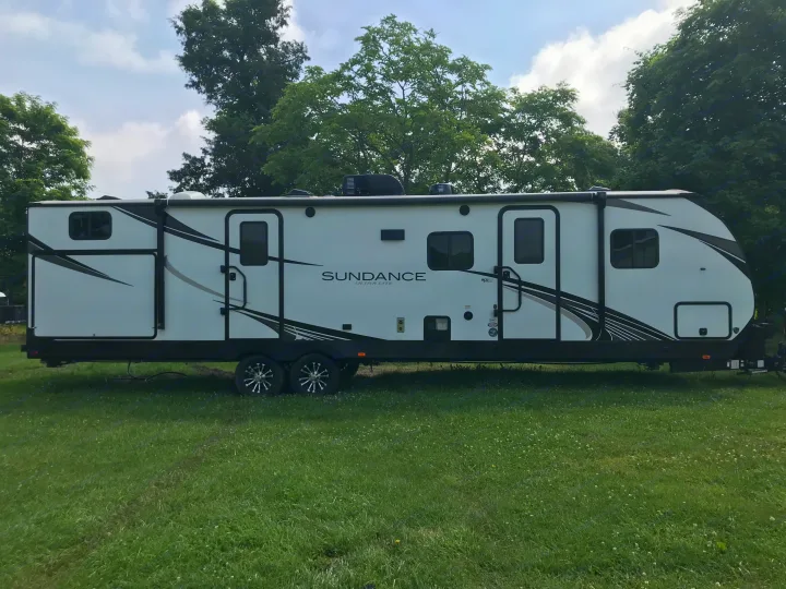 White Sundance Ultra-Lite travel trailer (RV rental) parked on green grass, surrounded by trees under a cloudy sky.. Heartland RVs Sundance Ultra Lite 2021