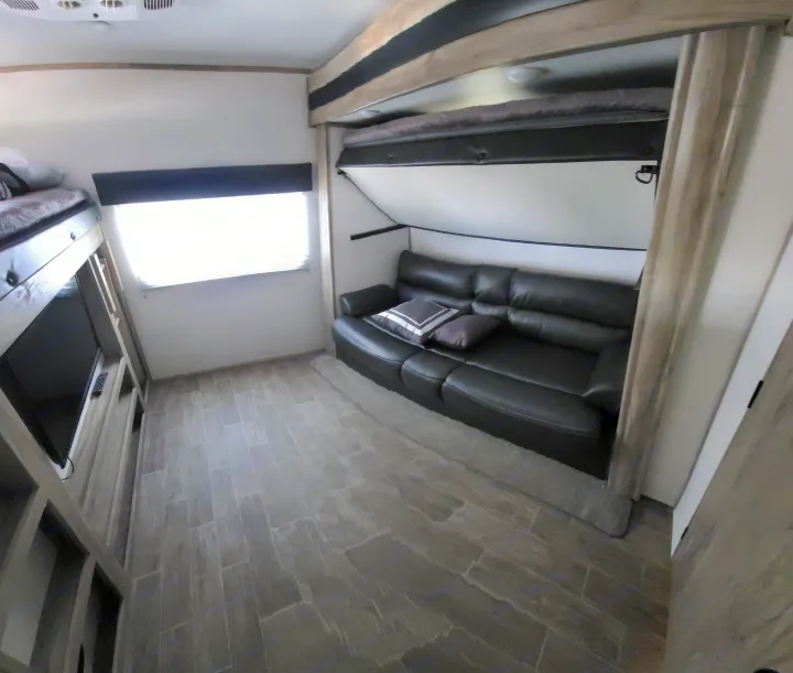 RV rental interior featuring a black leather sofa, pull-down upper bunk, bunk beds with TV, and wood-look tile flooring.. Forest River Cherokee Alpha Wolf 2021