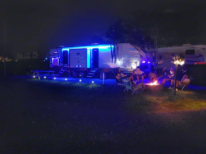 An RV rental campsite at night with friends gathered around a campfire by a brightly blue-lit RV and picnic table.. Forest River Cherokee Alpha Wolf 2021