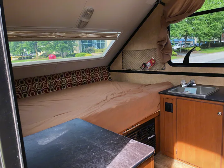Interior of an RV rental showing a made bed, compact sink, and Dometic unit, with large windows overlooking trees and buildings.. Starcraft Comet 2015