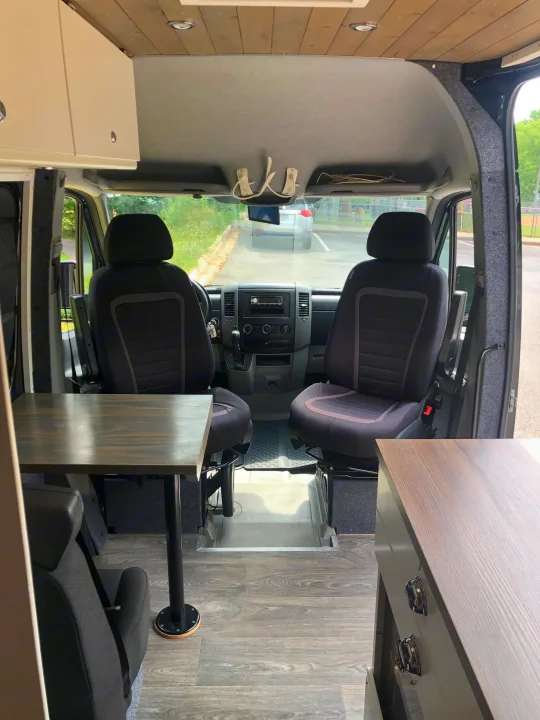 RV rental camper van interior showing front cabin seats, dashboard, a table, and overhead storage, with a wooden ceiling.. Mercedes Sprinter 2012