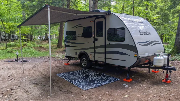 This RV rental is set up at a wooded campsite with an awning, rug, and leveling gear, perfect for outdoor adventures.. Prolite RV Evasion 16 2022