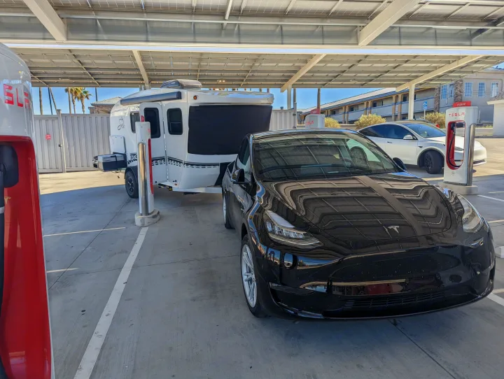 A black Tesla Model Y towing a sleek rv rental trailer charges at a sunny Supercharger station under solar panels. EV adventure!. Intech Sol Horizon Rover 2022
