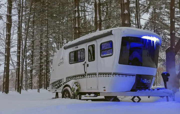 This white travel trailer RV rental, glowing blue with icicles, is set in a peaceful snowy forest, ideal for winter camping.. Intech Sol Horizon Rover 2022
