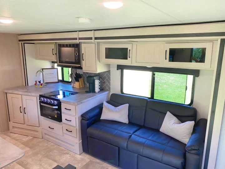 Modern RV rental interior: well-equipped kitchen with stove, microwave & sink. Comfortable black sofa by bright windows.. Forest River two bedroom Apex ultralite 2022